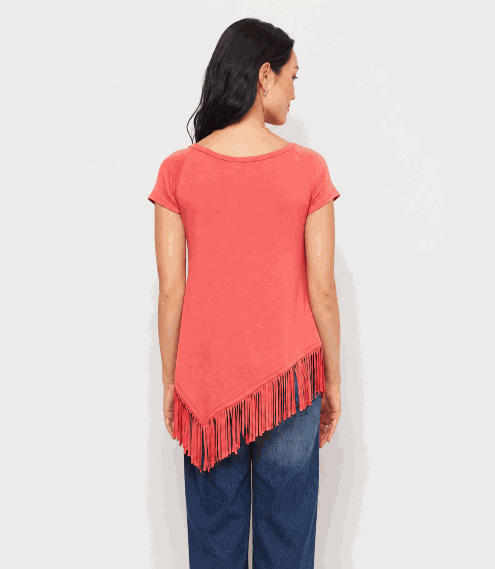 A woman shown from the back wearing a coral pink short-sleeve top with fringe trim along the asymmetrical hem, paired with blue jeans, against a plain light gray background.