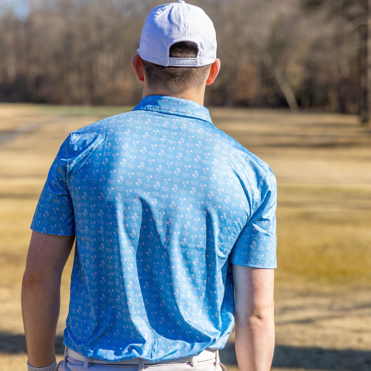 Man wearing a blue patterned shirt and white cap standing in a field with trees in the background