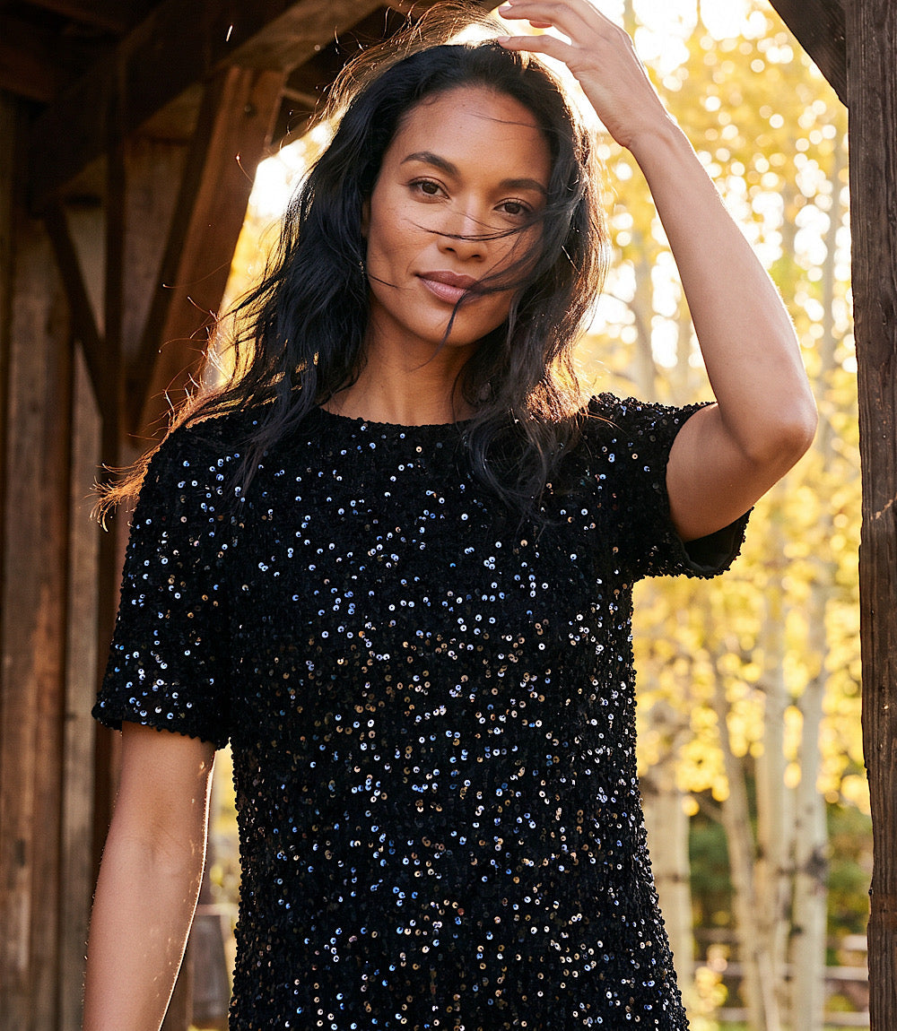 A woman wearing a short-sleeve black sequined top or dress, posing outdoors under a wooden structure with bright, sunlit trees in the background.