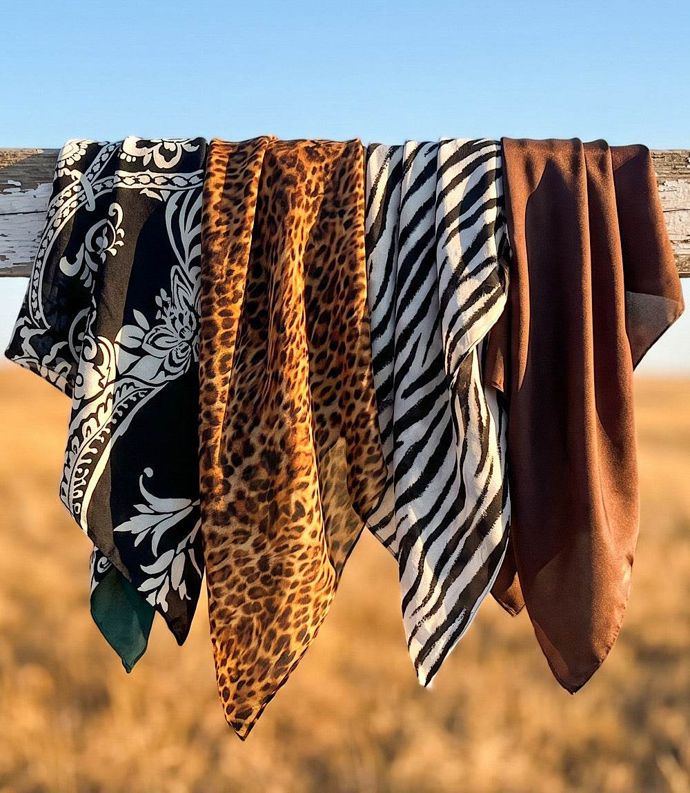 Four patterned square scarves in different designs are draped over a rustic wooden fence outdoors, with a blurred field and blue sky in the background.