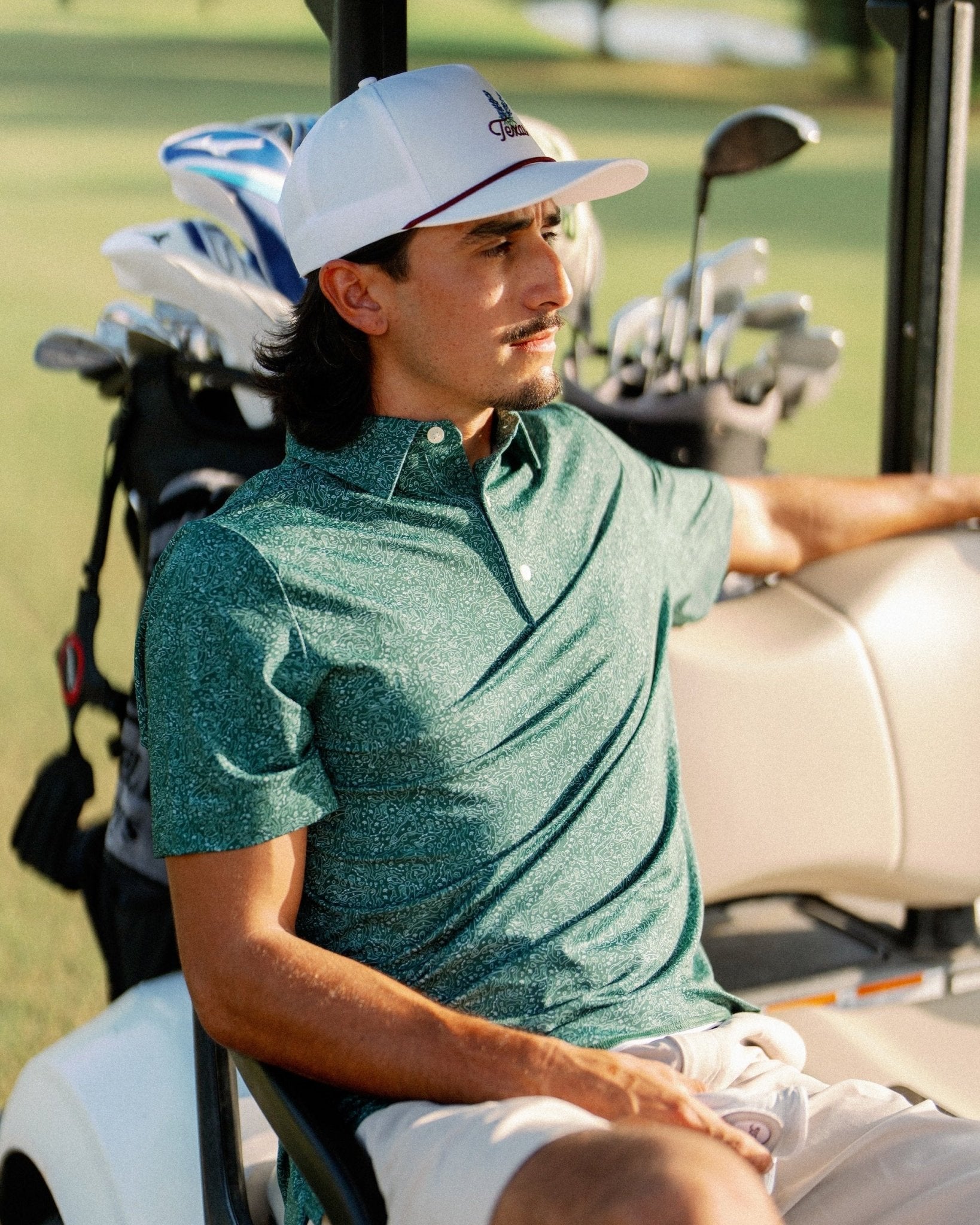 A man wearing a green heathered short-sleeve polo shirt and white shorts sits in a white golf cart on a golf course, wearing a white baseball cap.