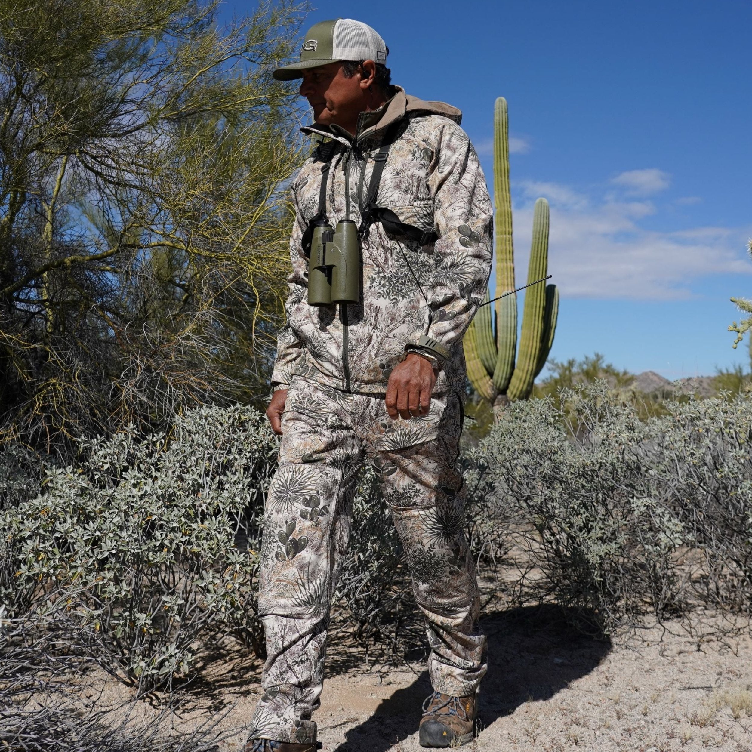 Person in camouflage gear standing in a desert landscape with cacti and shrubs.