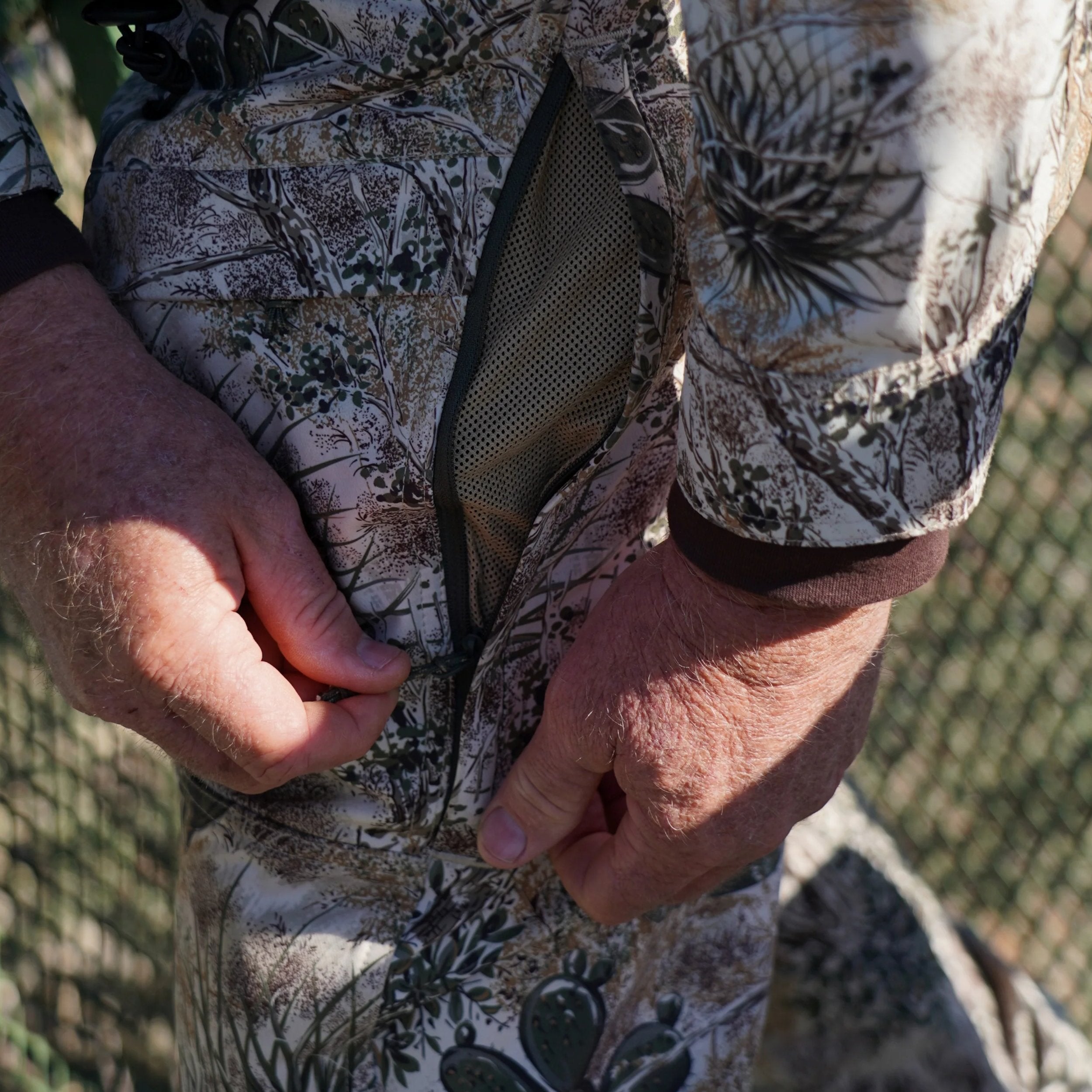 Close-up of hands buttoning a camouflage jacket with a chain-link fence in the background.