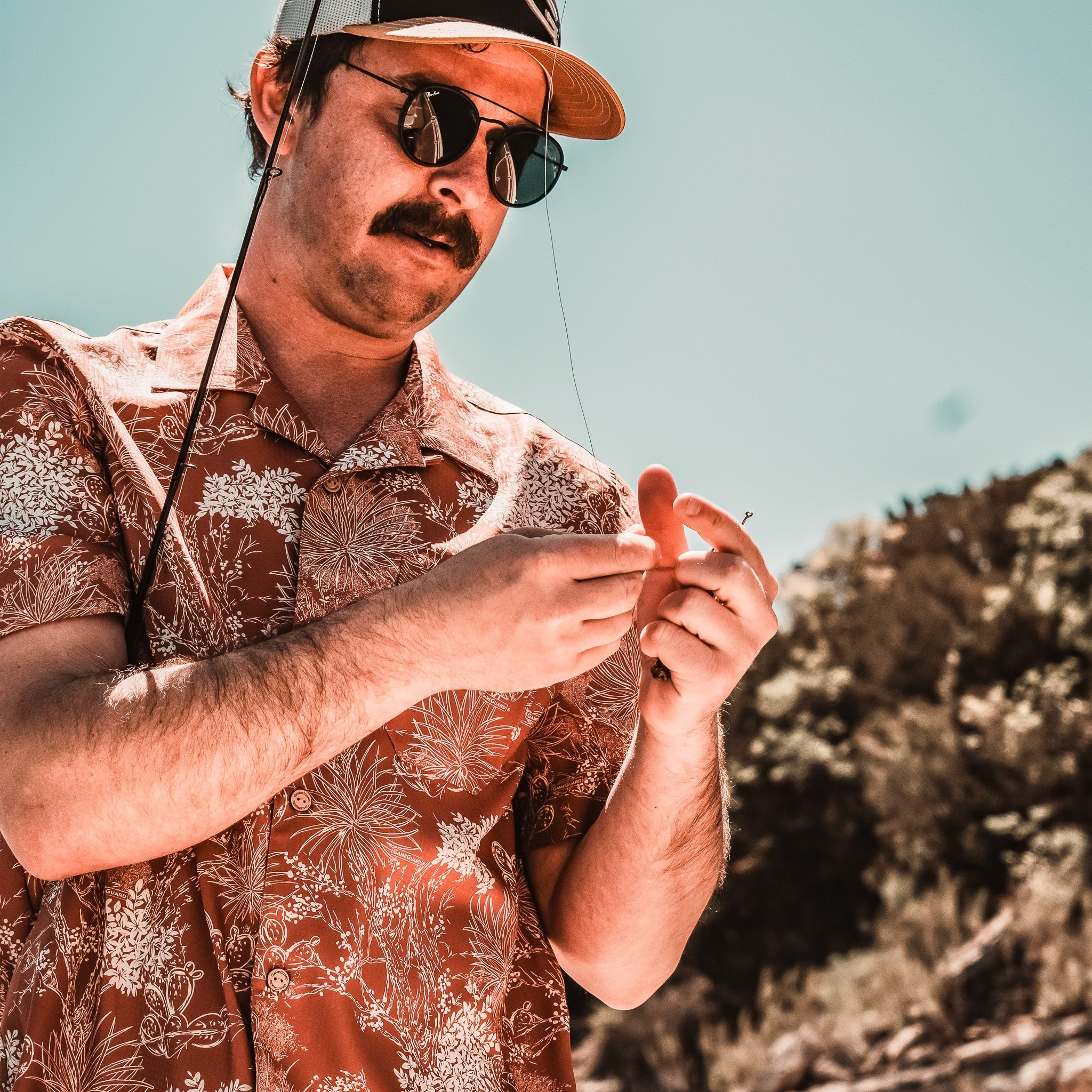 Man in a patterned shirt and cap fishing outdoors with trees in the background
