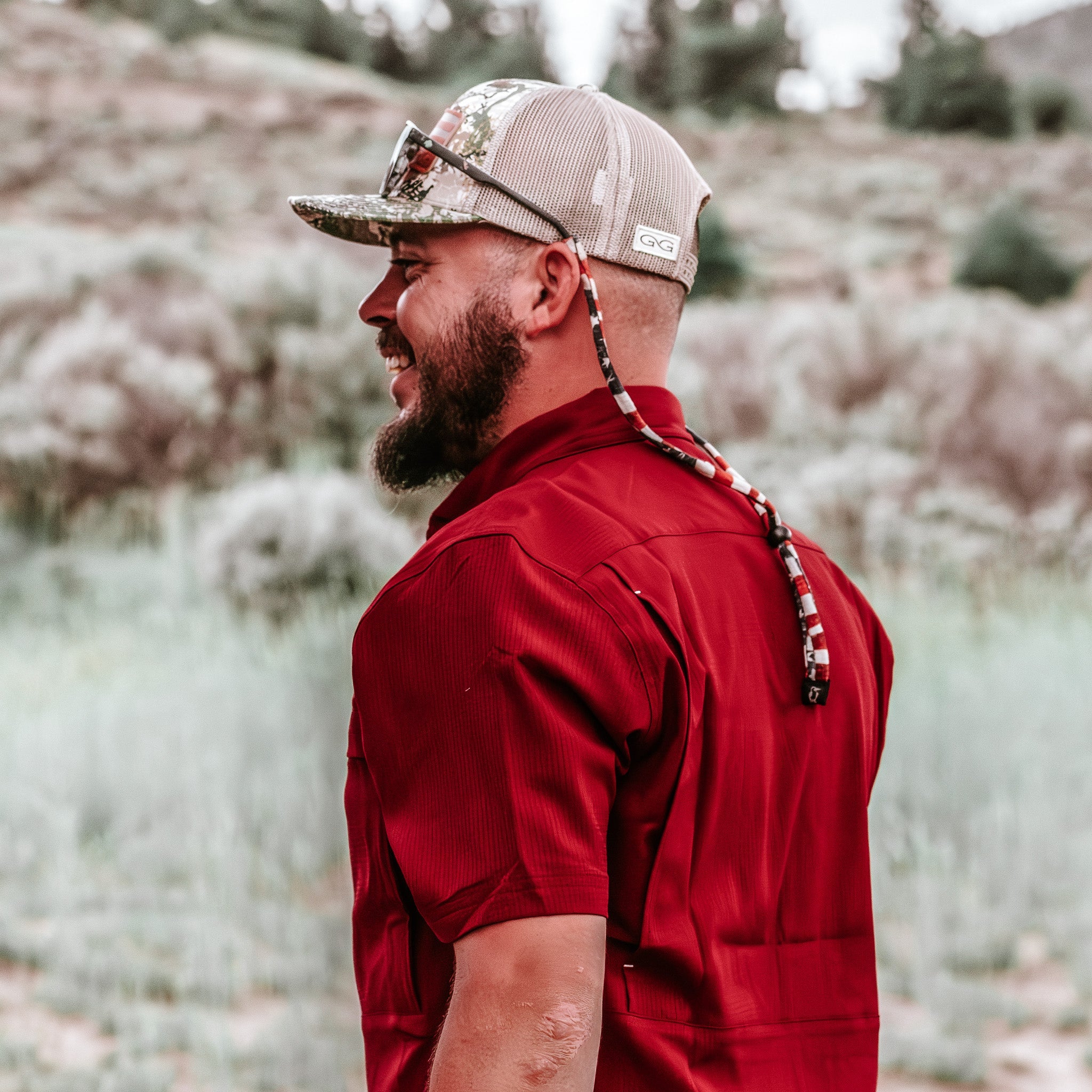 Man wearing a red shirt and camouflage cap with a bow on his head, standing outdoors.