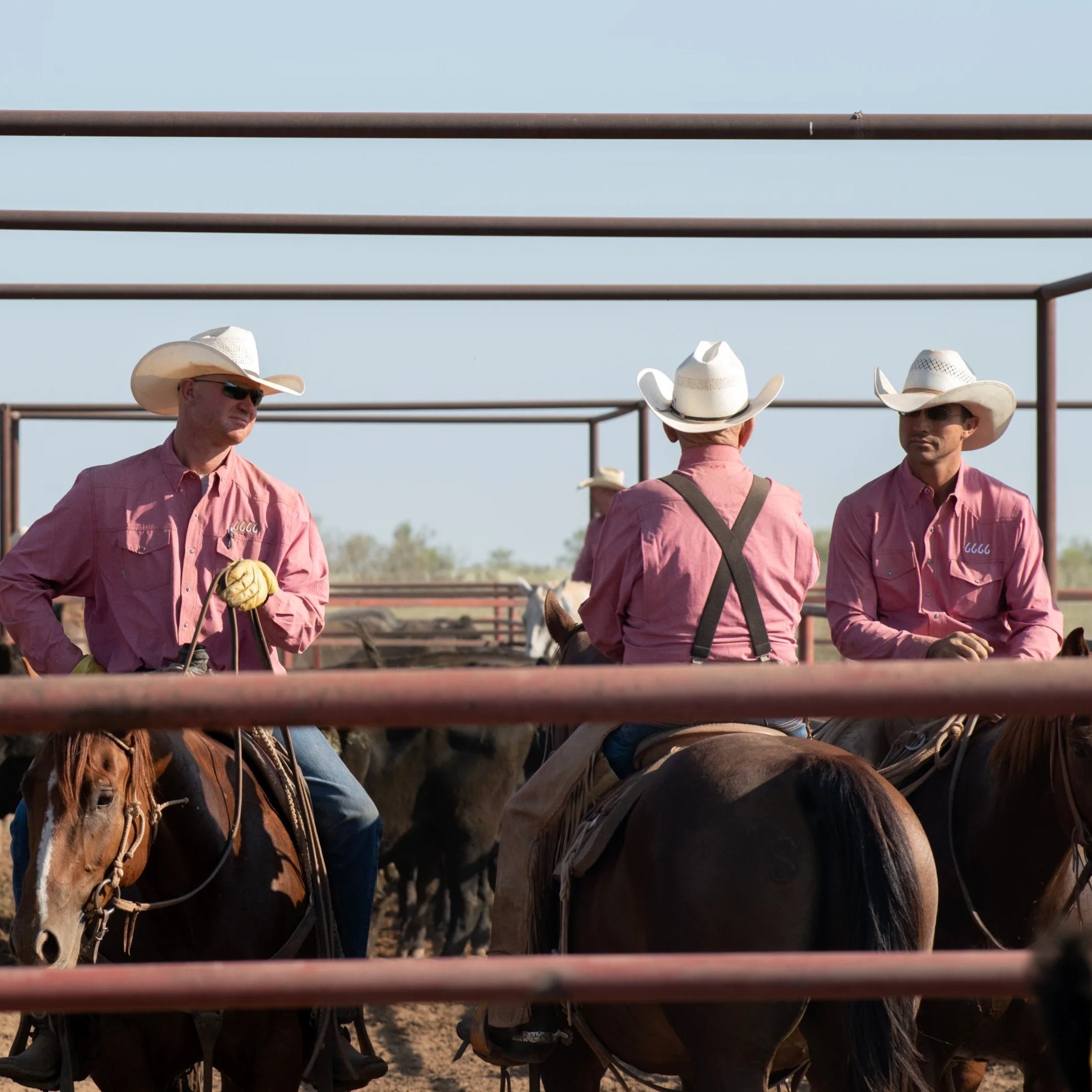 Three cowboys on horseback in a pen wearing pink shirts and white hats.