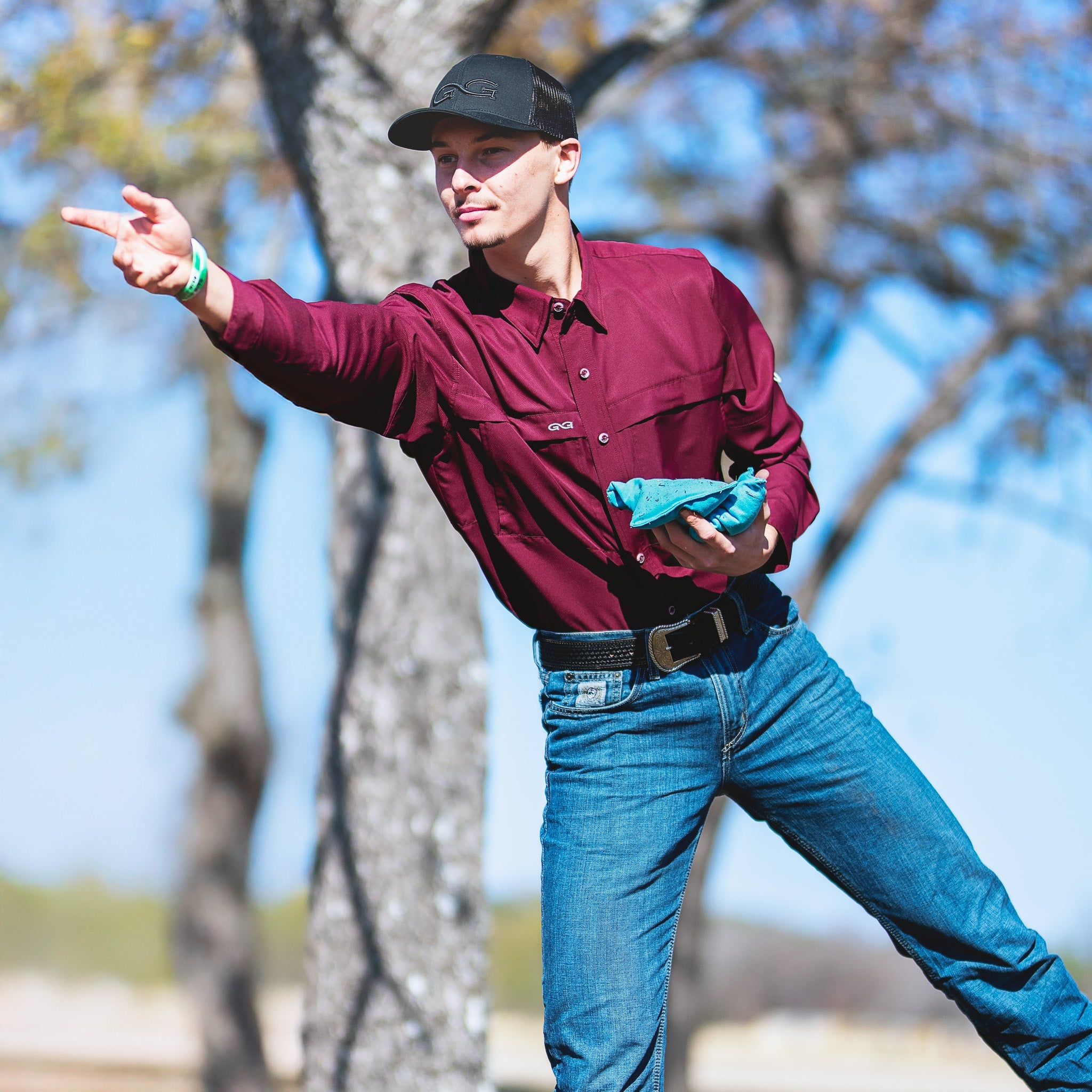 Man in maroon shirt and blue jeans holding a blue object outdoors with trees in the background