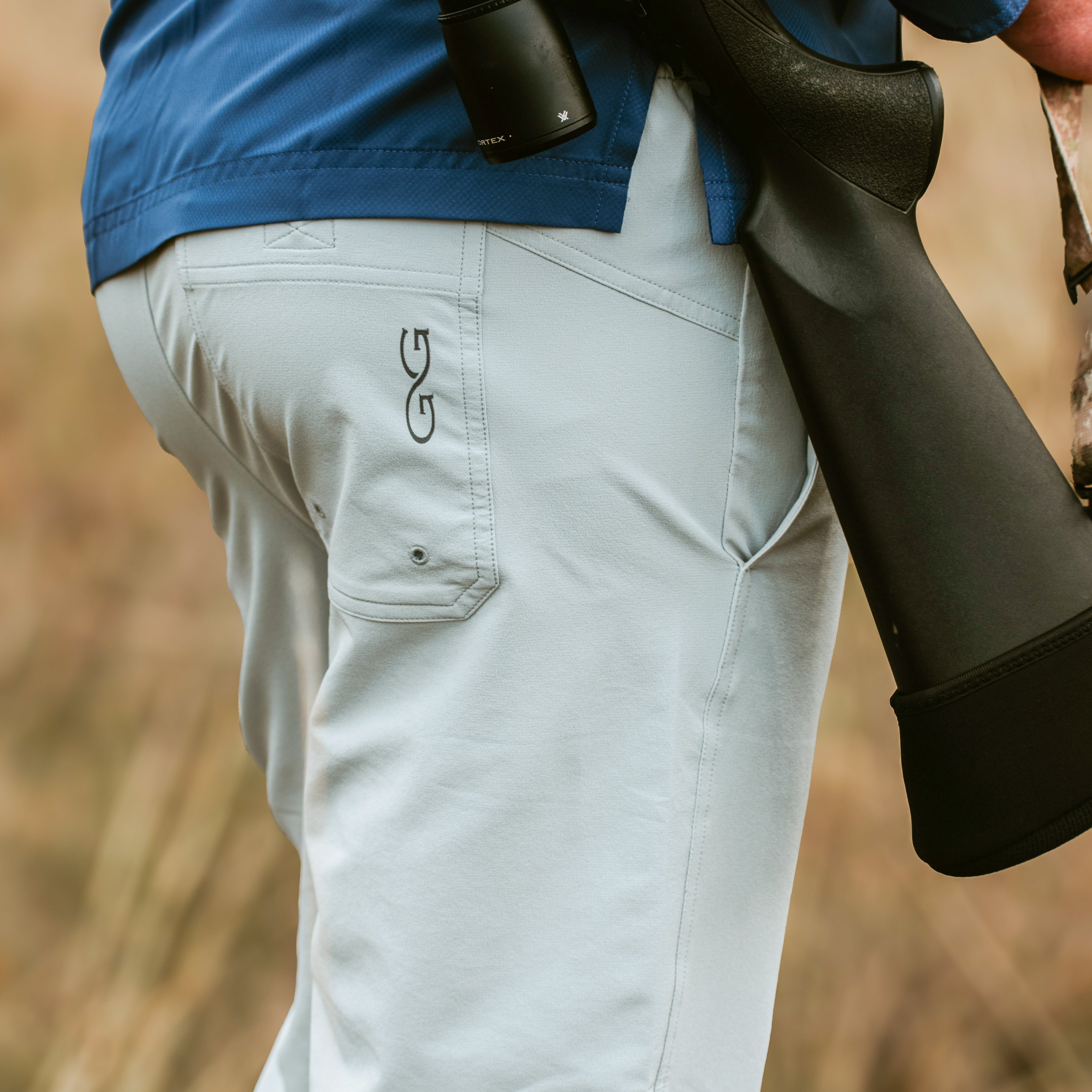 Person wearing light gray pants with a brand logo, holding a rifle in an outdoor setting.
