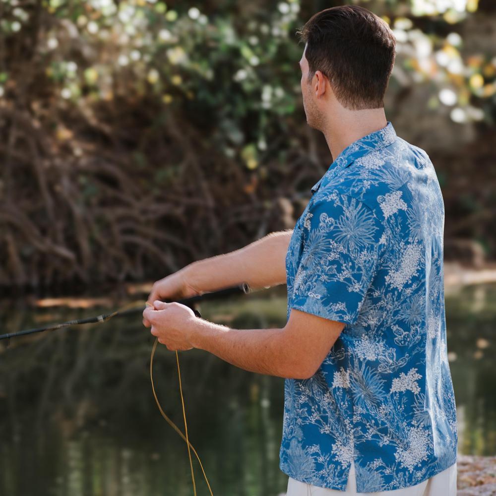 Man in a blue floral shirt fishing by a body of water with greenery in the background