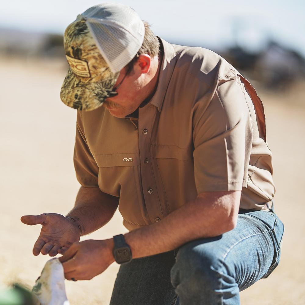 Man in outdoor setting wearing a cap and button-up shirt, holding a bag.