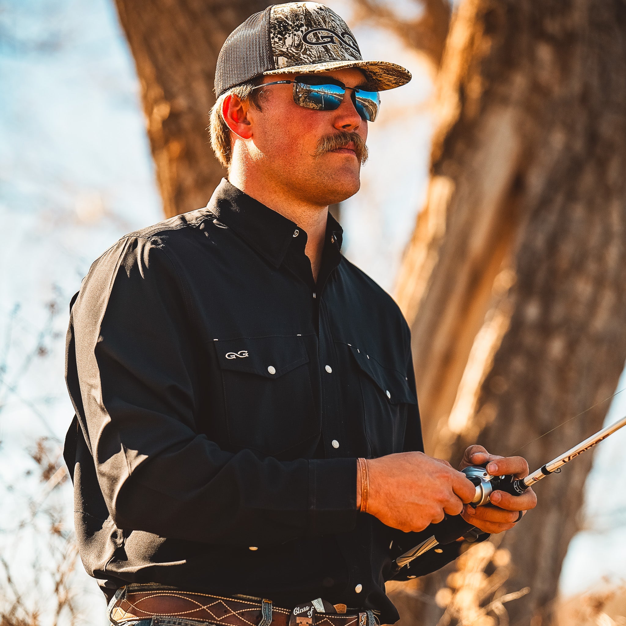 Man in outdoor setting holding a bow and arrow, wearing a black shirt and camouflage cap.