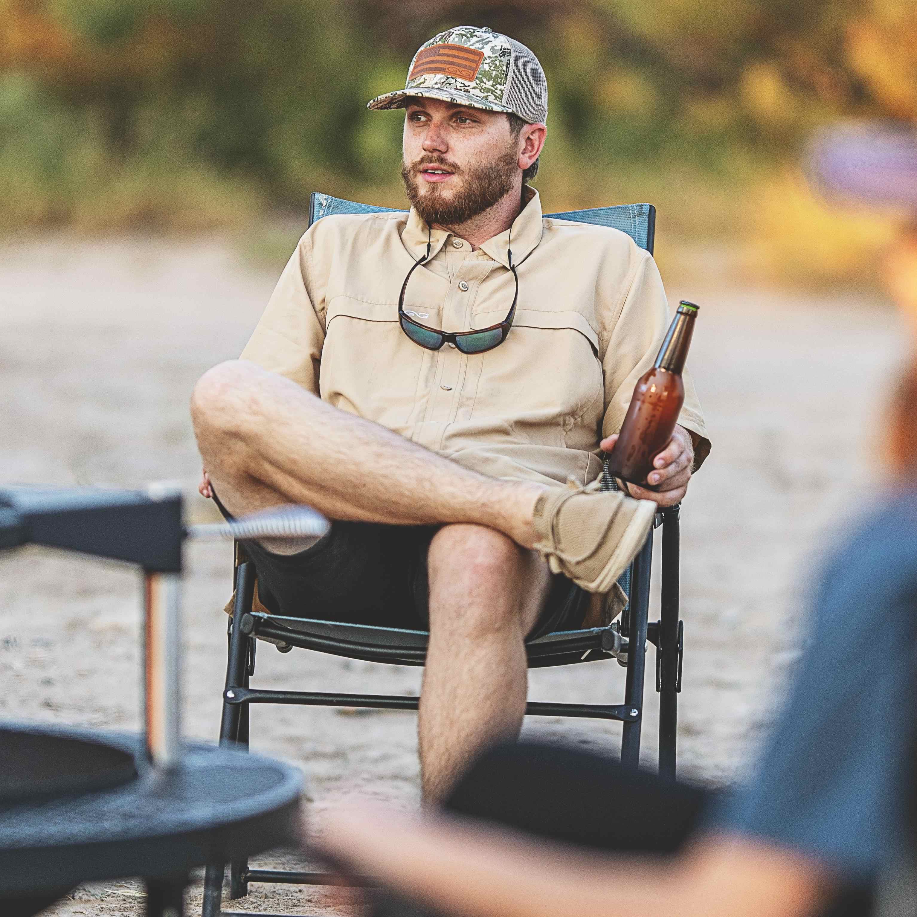 Man sitting outdoors in a chair holding a beer bottle, wearing a cap and sunglasses around his neck.