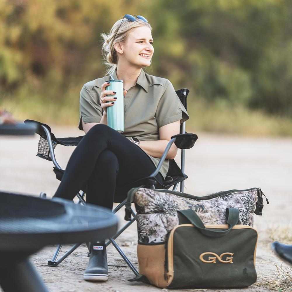 Woman sitting in a camping chair outdoors with a bag and cup nearby