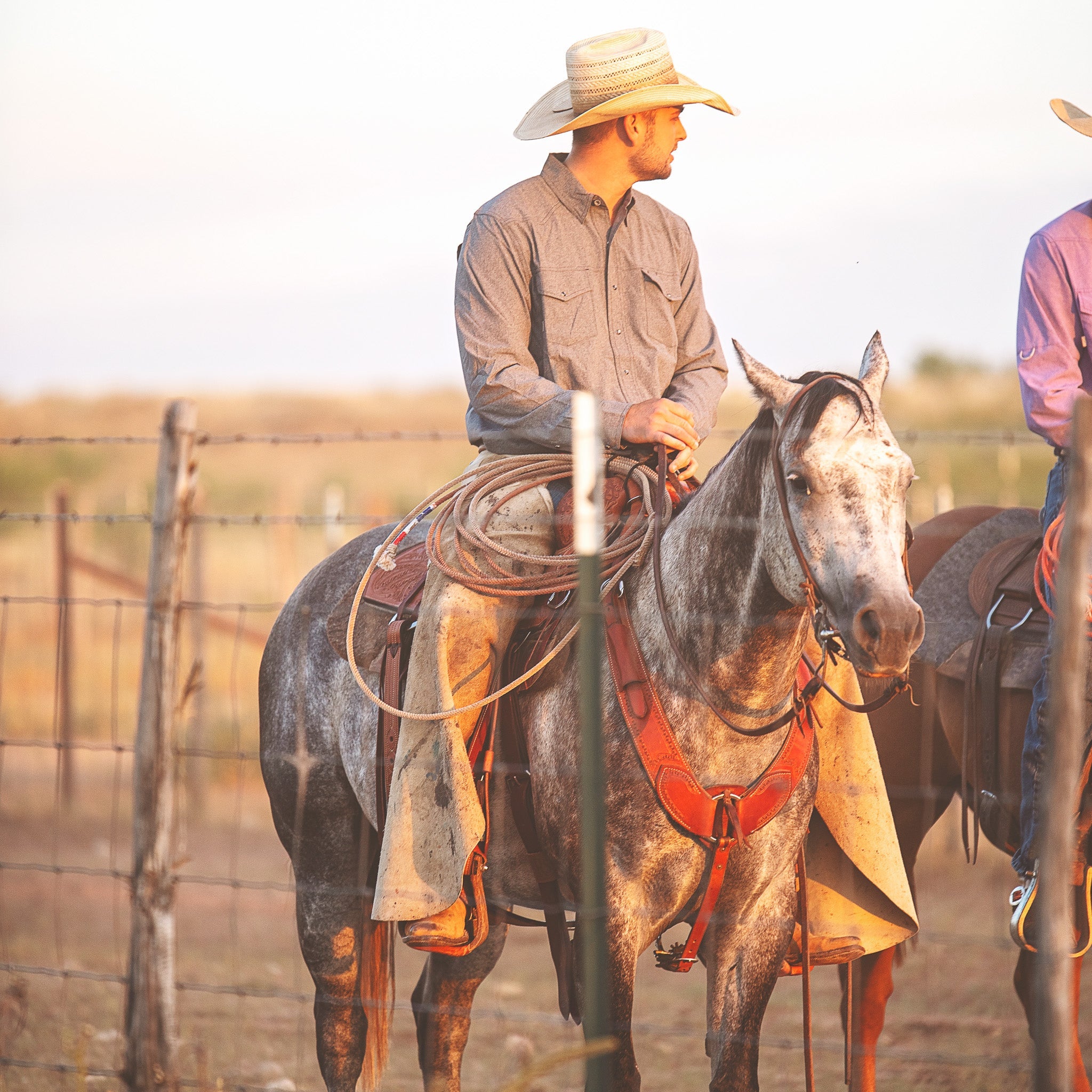 Two cowboys on horseback in a rural setting with a fence and open field.