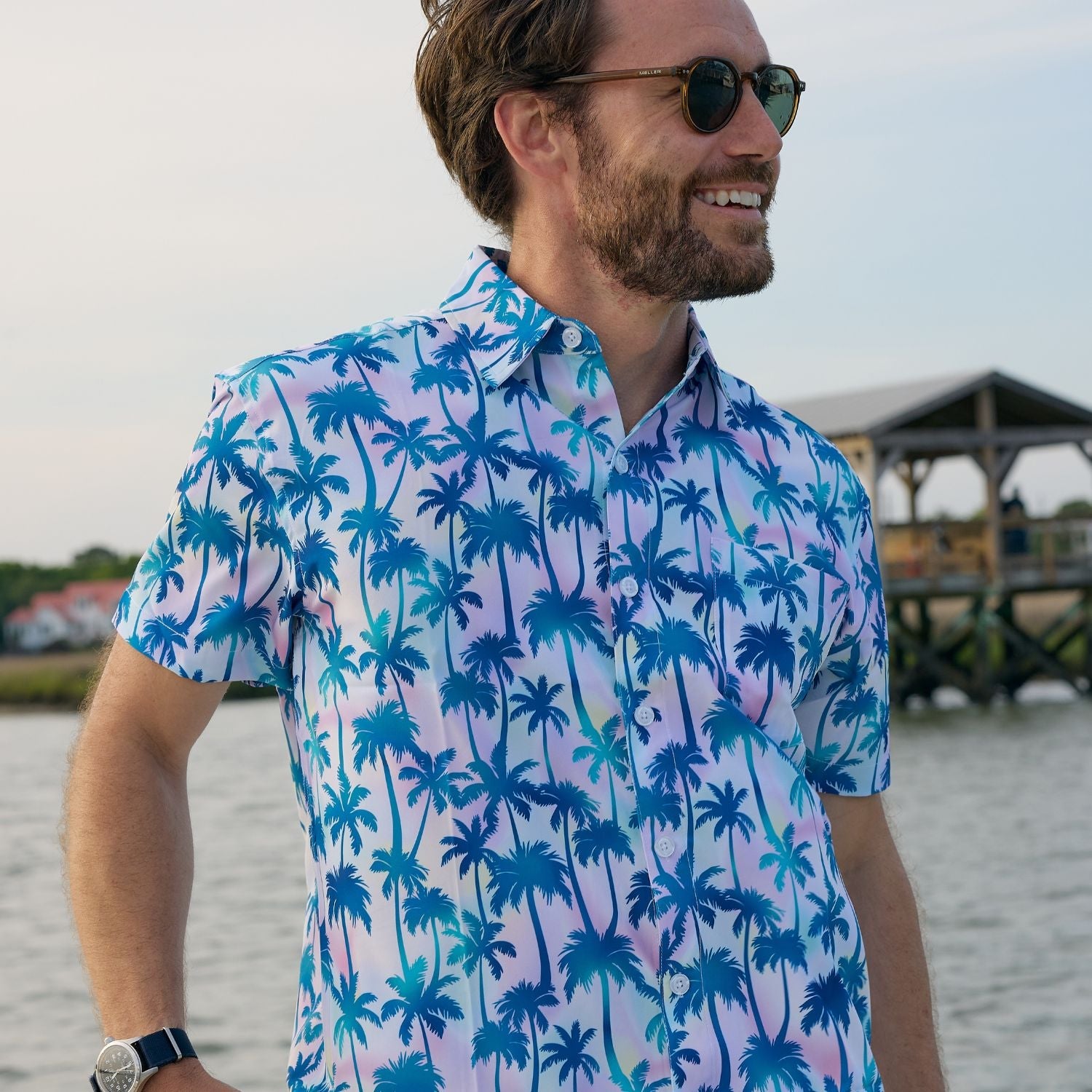 Man wearing a colorful palm tree patterned shirt by a waterfront.