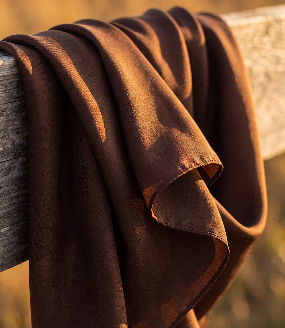 A draped brown fabric, likely a scarf or throw blanket, hanging over a rustic wooden fence post in warm golden sunlight.