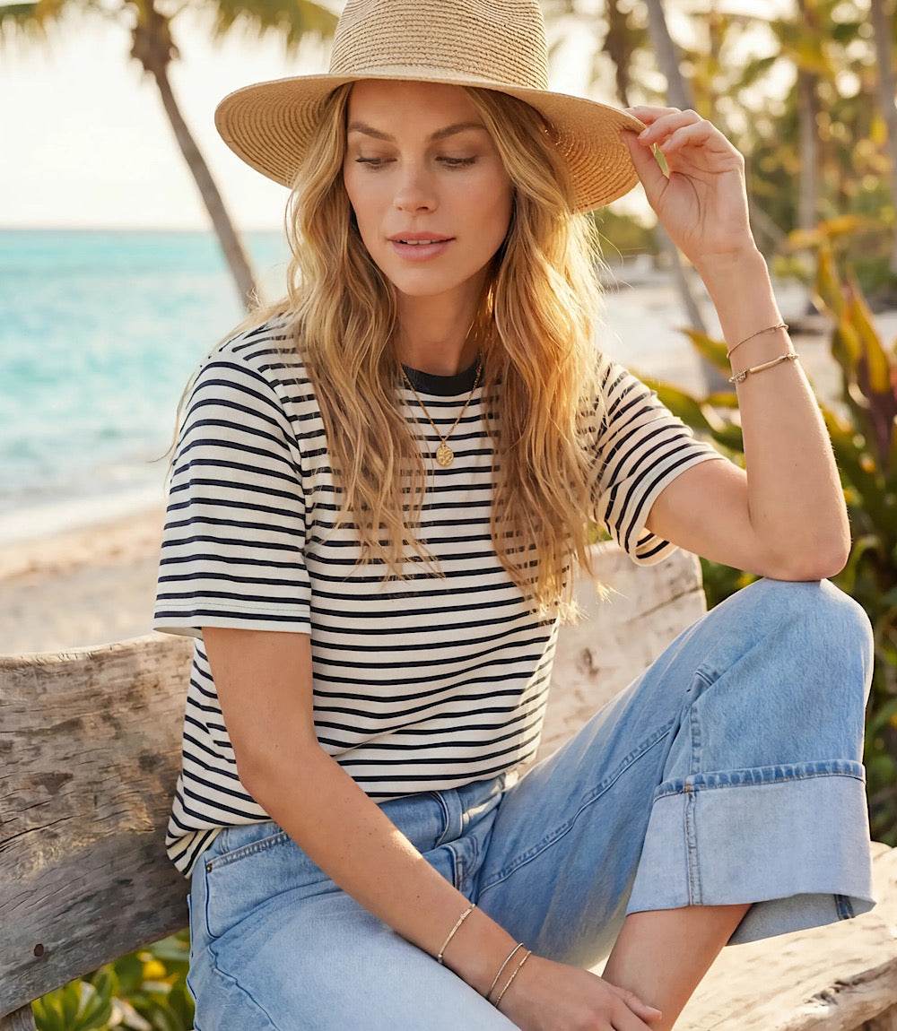 Woman sitting on a wooden bench by the beach wearing a striped shirt and wide-brimmed hat.