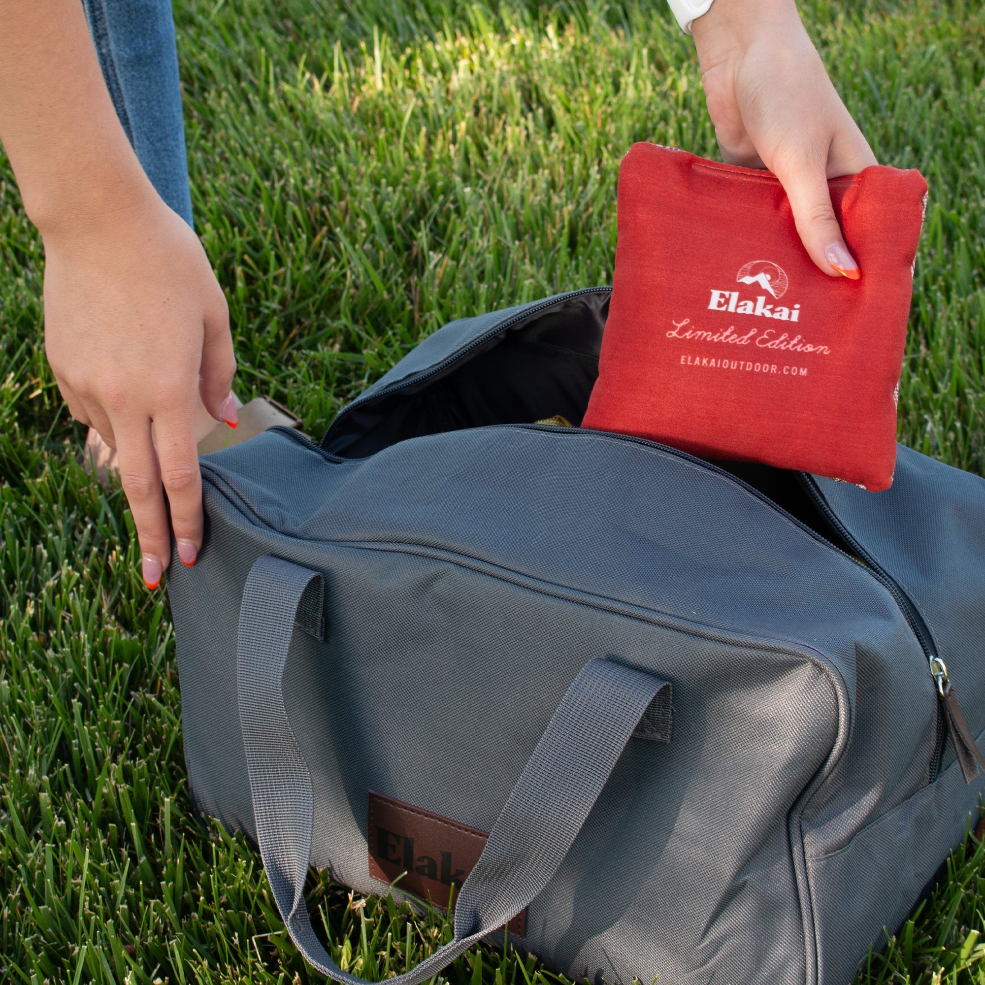Person placing a red Elakai pouch into a gray duffel bag on grass
