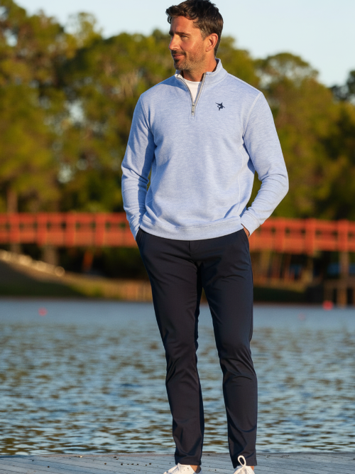 Man wearing a light blue sweater and dark pants standing by a body of water with trees in the background.