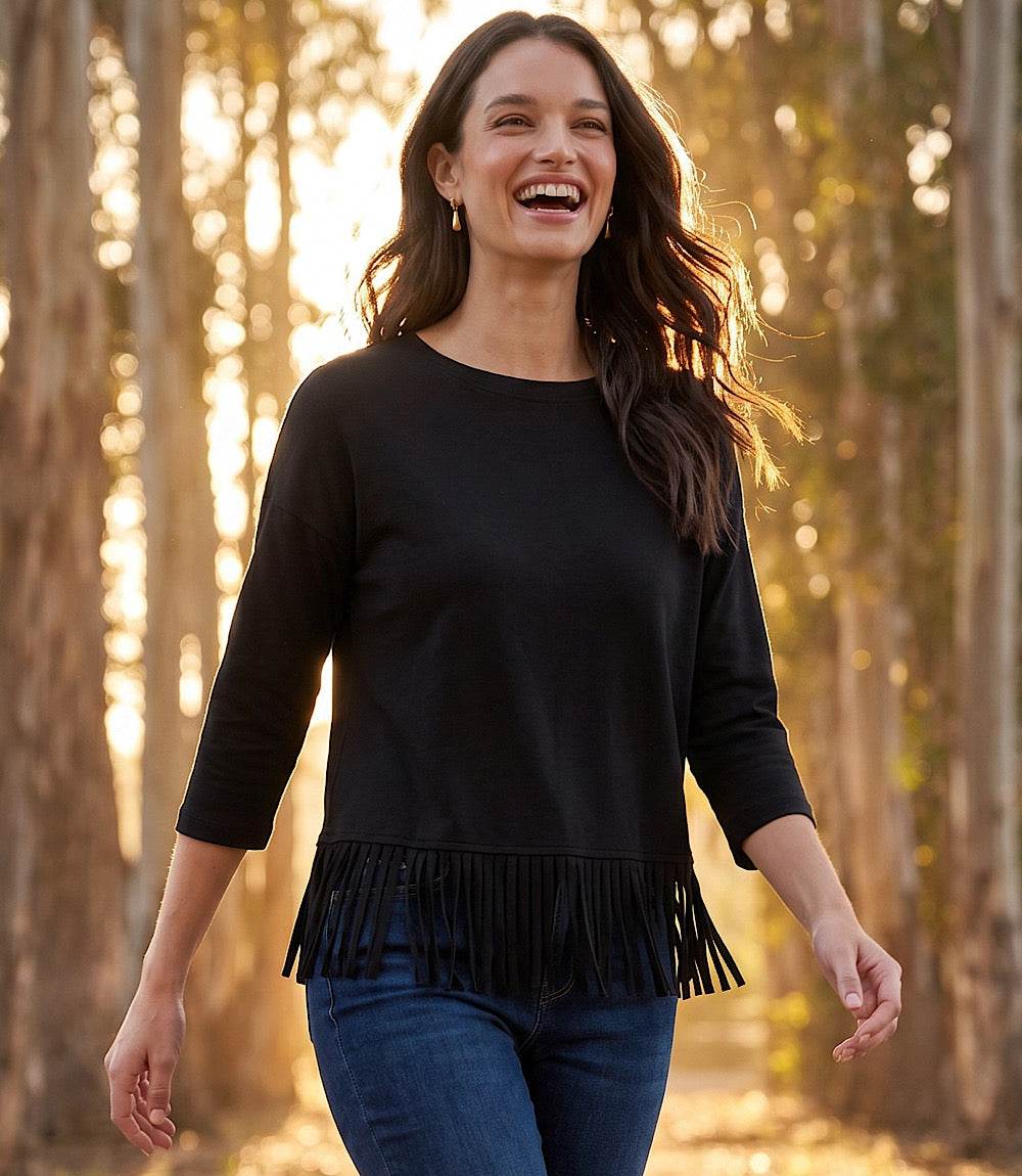 A woman smiling outdoors in a black three-quarter sleeve top with fringe trim at the hem, paired with blue jeans, set against a warm, sunlit forest background.
