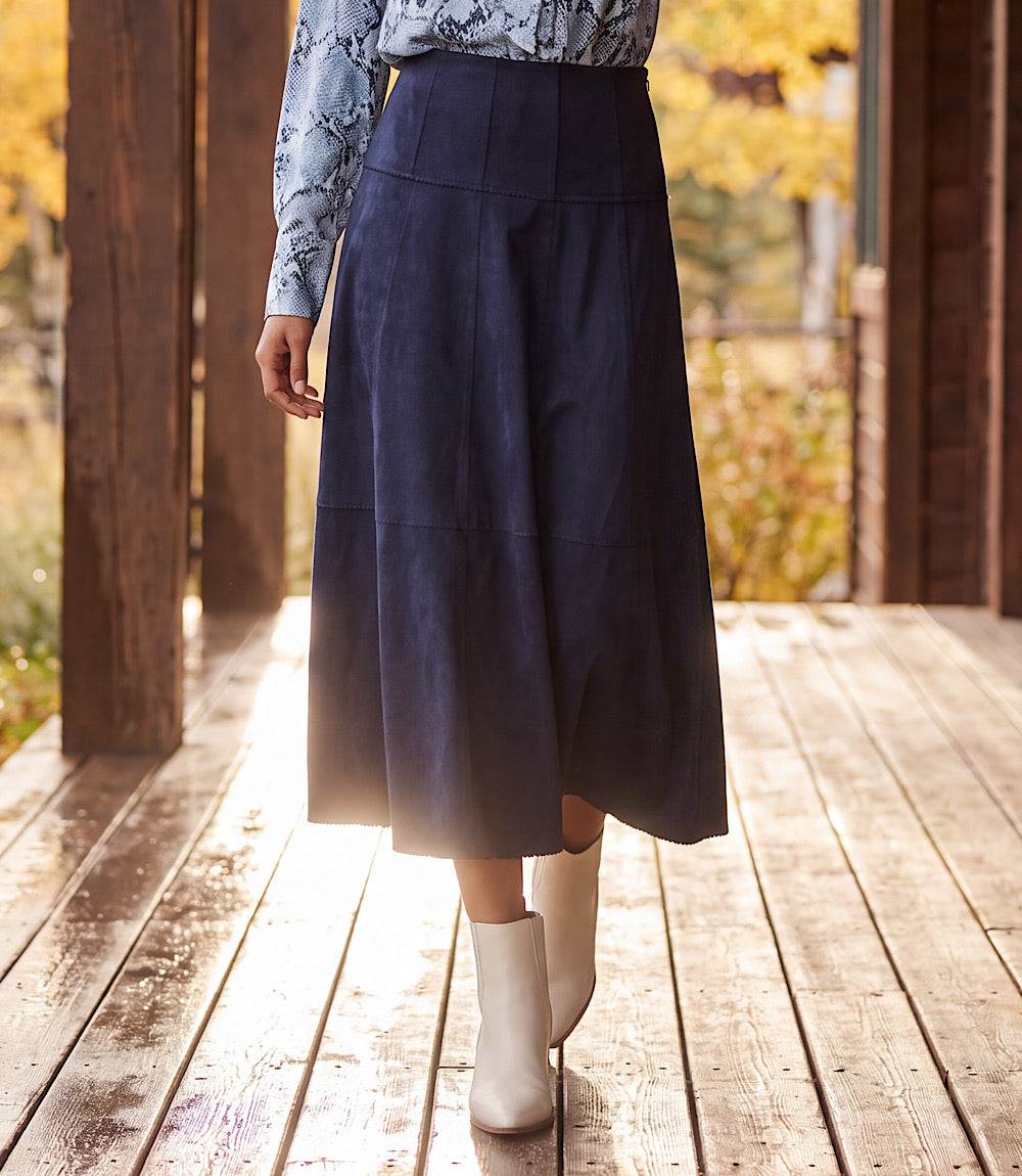 A person wearing a dark navy midi skirt, a light blue patterned long-sleeve top, and white ankle boots, standing on a wooden deck with a blurred outdoor background of autumn trees.