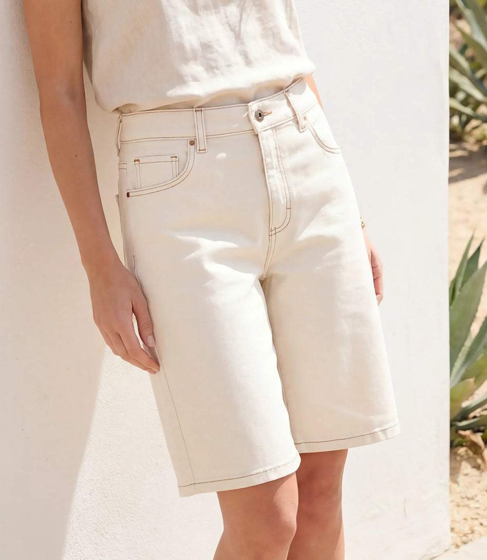 A person wearing off-white denim shorts and a matching off-white top, leaning against a white wall in a sunlit setting with a plant visible at the edge.