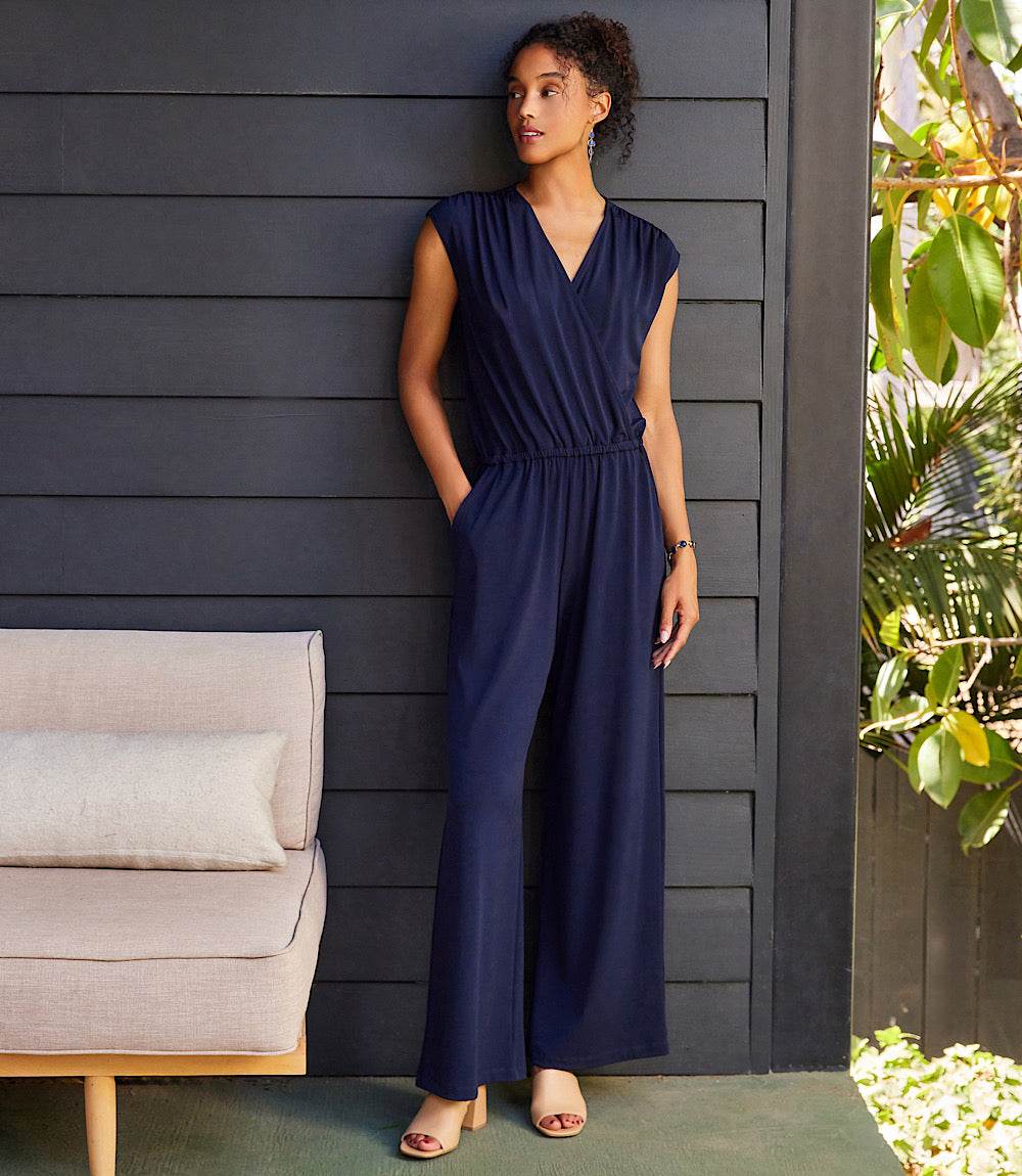A woman wearing a navy blue sleeveless wrap-style jumpsuit with wide legs, standing against a dark gray wooden wall next to a light beige sofa and green plants.