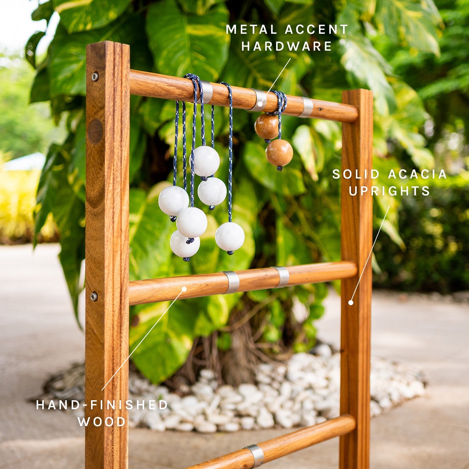 A wooden garden trellis or ladder-style stake with labeled callouts pointing to metal accent hardware, solid acacia uprights, and hand-finished wood. The product is shown outdoors with green foliage and a stone path in the background.