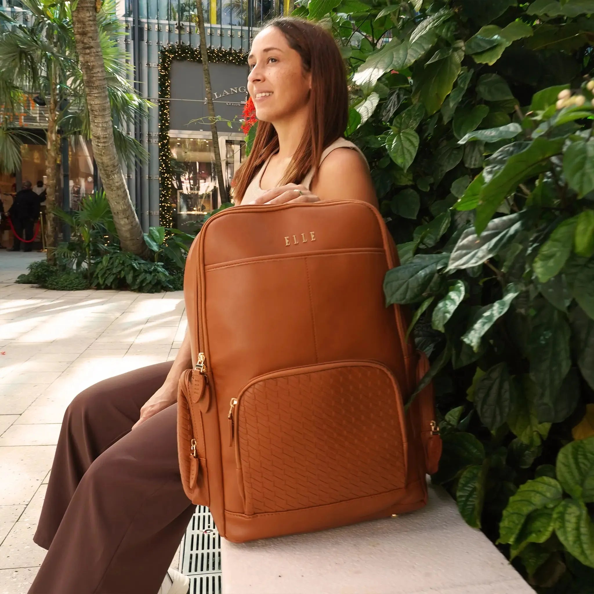 Woman holding a brown backpack with 'ELLE' branding in an outdoor setting.