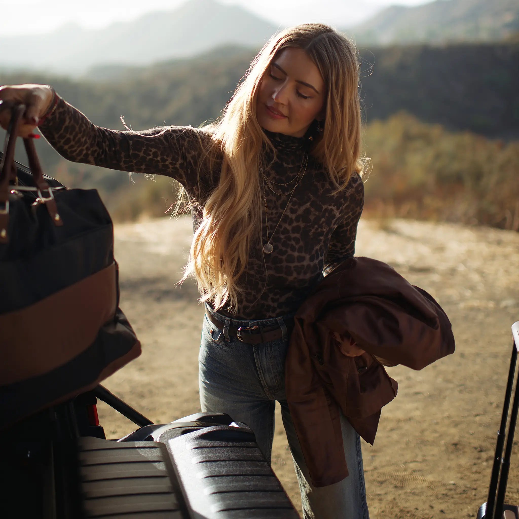 Woman with a leopard print sweater and jeans standing next to a vehicle in a natural setting.