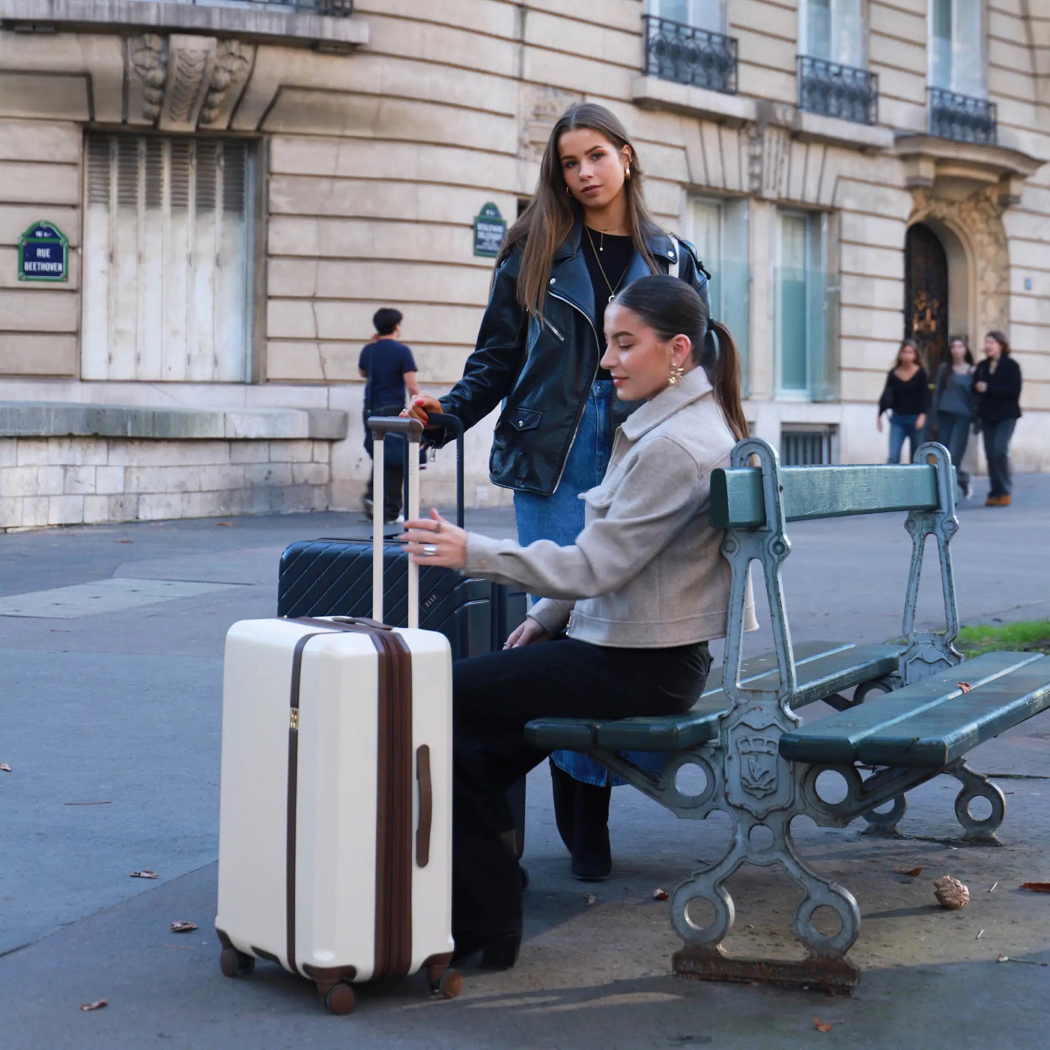 Two women with luggage on a city street, one sitting on a bench.