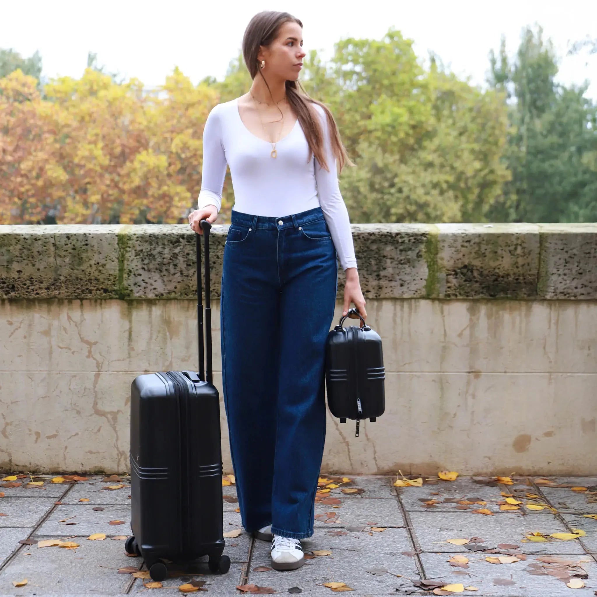 Woman holding a suitcase and a small bag in an outdoor setting with trees in the background