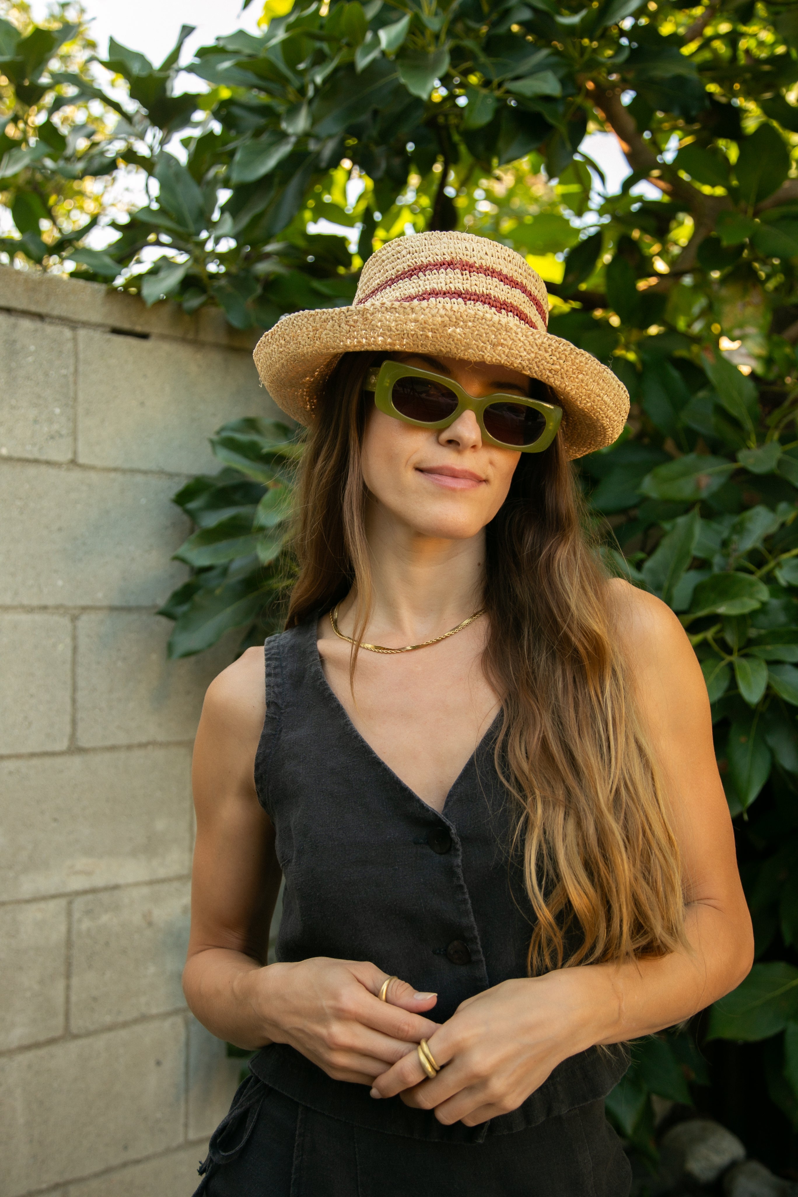 Woman wearing a straw hat and sunglasses standing in front of green foliage.