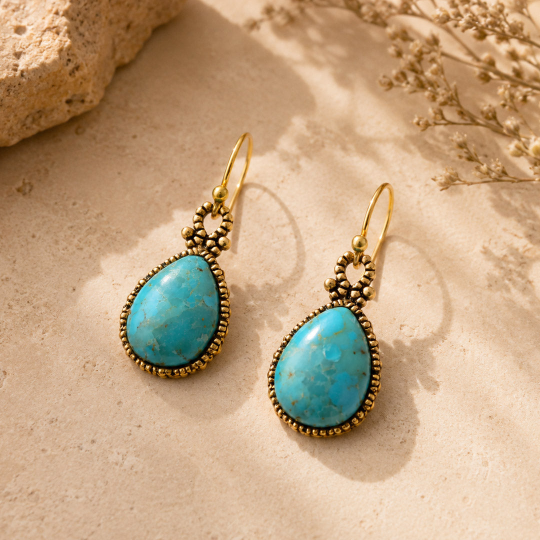 Turquoise teardrop earrings with gold accents on a sandy surface.