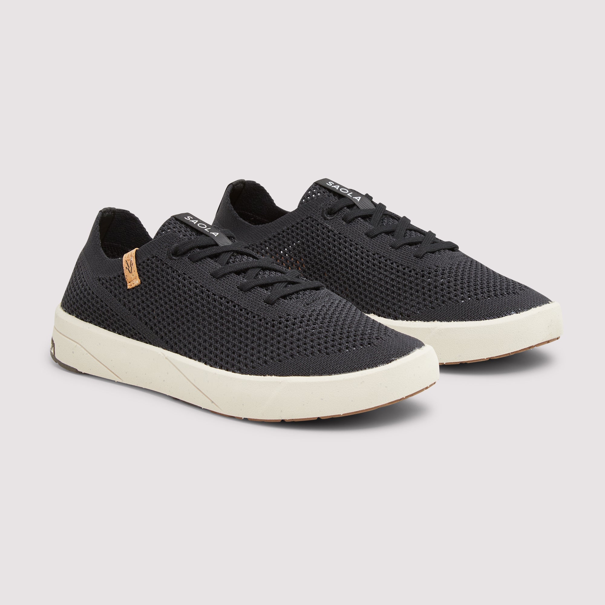 A pair of black mesh low-top sneakers with off-white soles and a small tan brand tag on the side, displayed against a light gray background.
