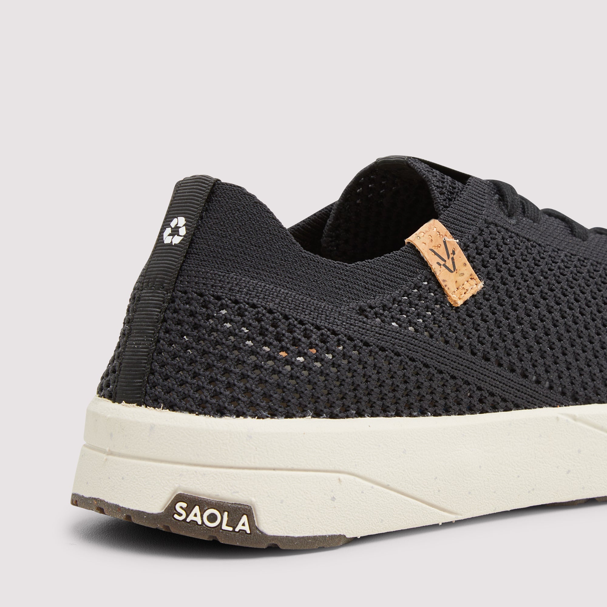 Close-up of a black knit sneaker showing the heel, side knit texture, a small tan leather logo tab, and a thick off-white sole with a brown SAOLA brand patch.