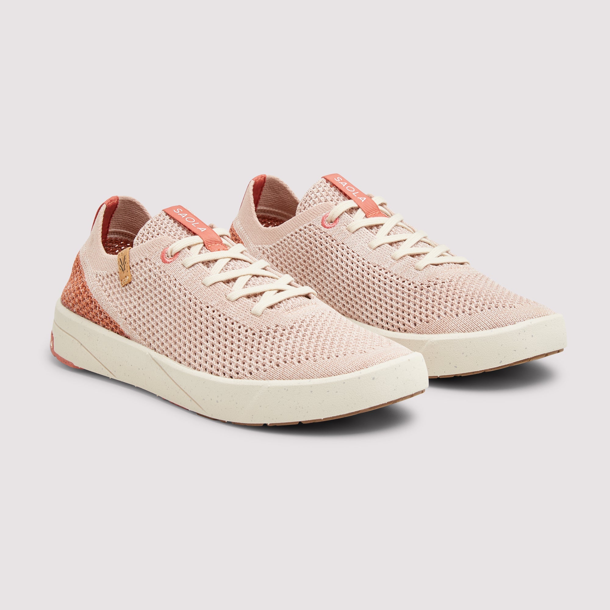 A pair of women's knit sneakers in a light pink and coral colorway, featuring off-white laces and thick cream-colored soles, shown against a neutral light gray background.