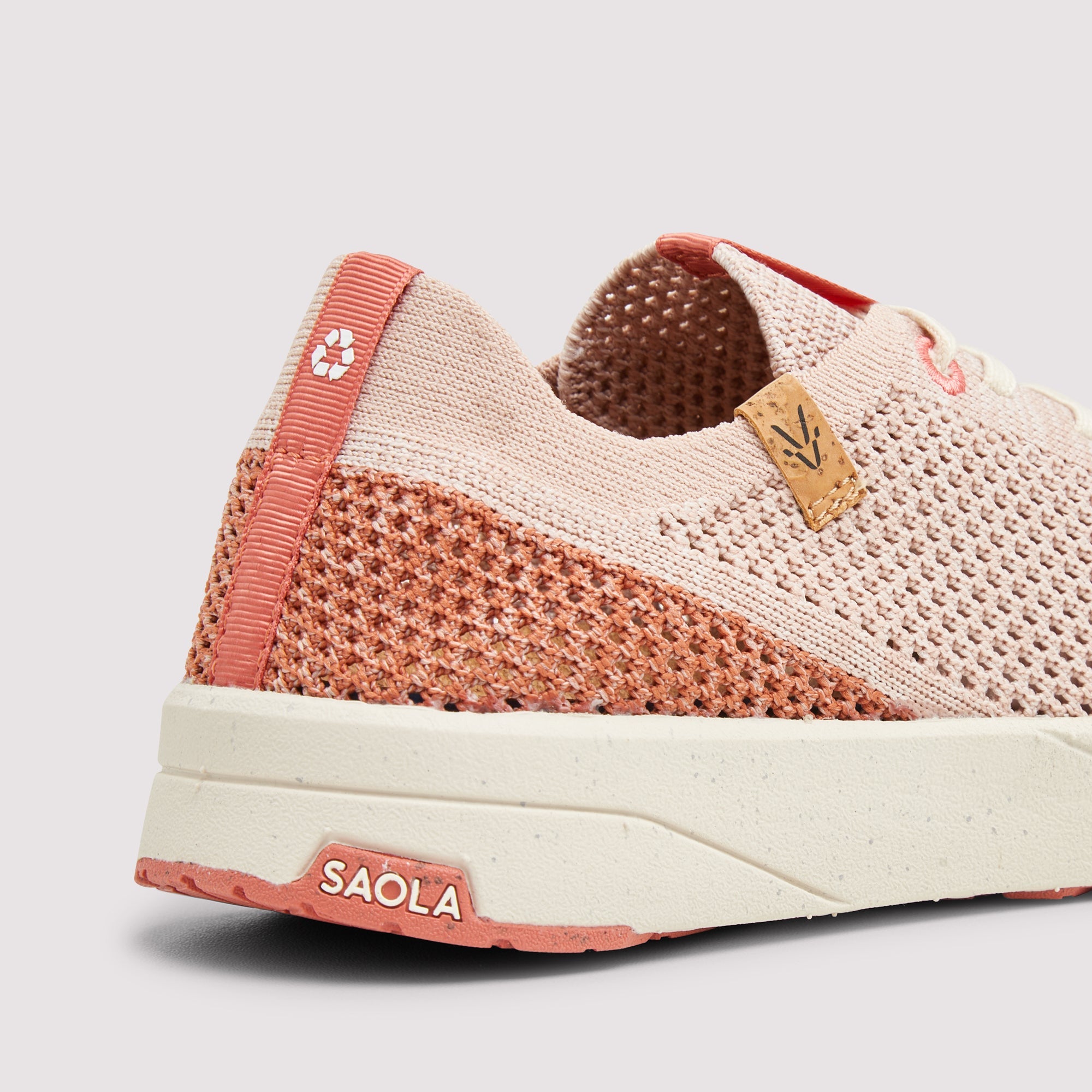 A close-up side view of a knit slip-on sneaker in light pink and coral pink, featuring a thick cream-colored midsole and a small tan leather logo tab on the side.