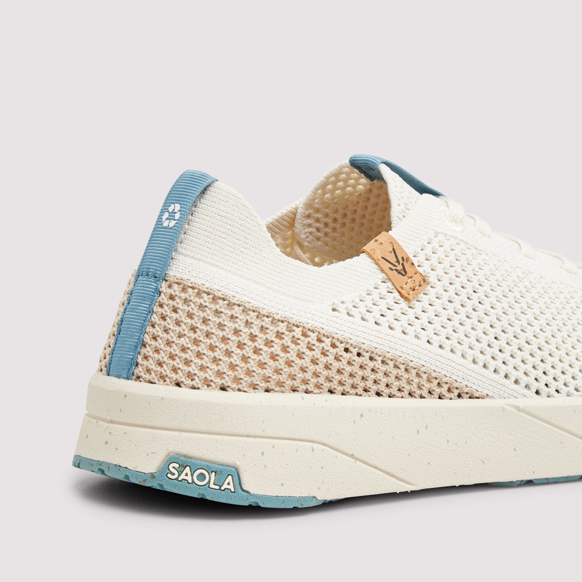 Close-up of a low-top athletic sneaker by SAOLA, showing the side profile. The shoe features a white knit upper, a beige mesh panel, a light blue heel tab, and a speckled off-white sole with a light blue 'SAOLA' logo on the heel.