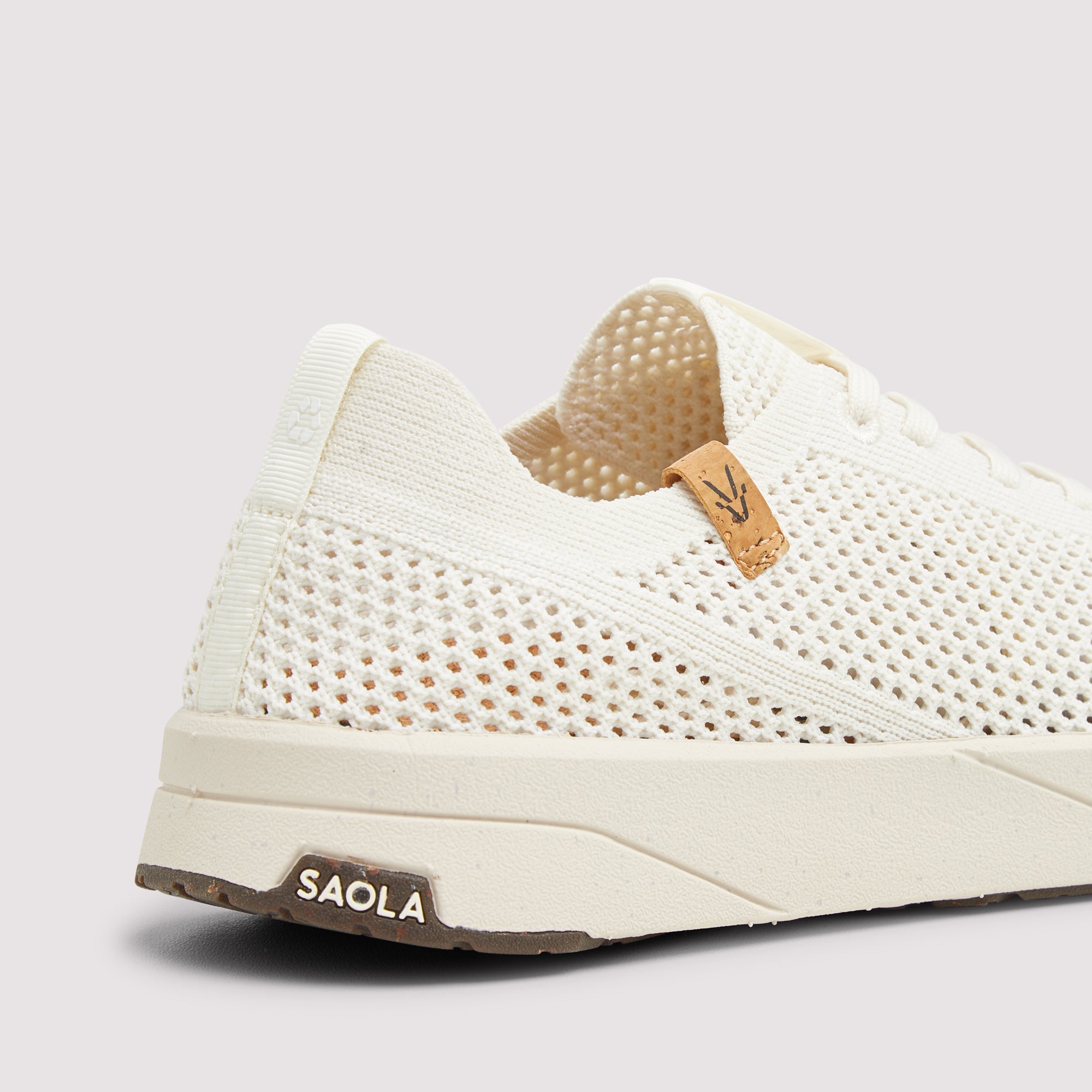 A close-up side view of a knit mesh sneaker in off-white with a light brown leather heel tab and a brown 'SAOLA' logo on the midsole heel.