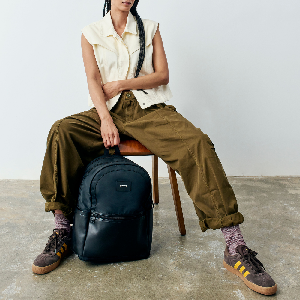 Person sitting on a chair with a black backpack, wearing a white sleeveless shirt, green pants, and brown shoes.