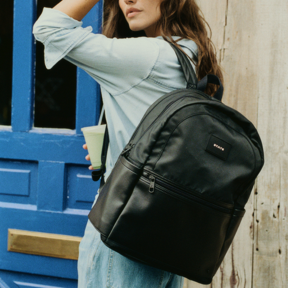 Person wearing a black backpack holding a drink against a blue door and wooden wall.