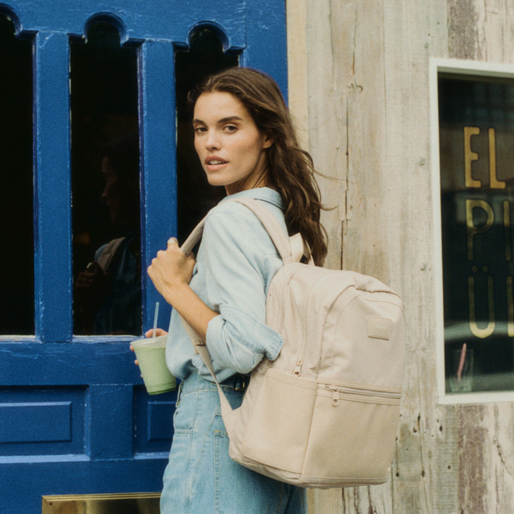 Woman with a beige backpack holding a drink, standing in front of a blue door and wooden wall.