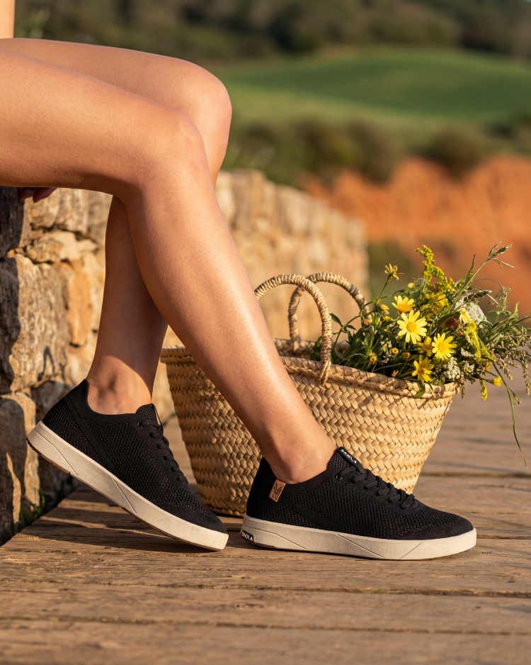 Person wearing black sneakers with white soles on a wooden floor, with a woven basket and flowers in the background.