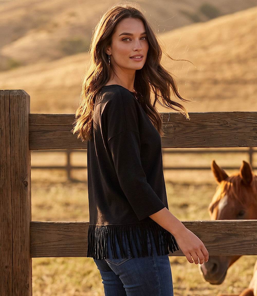 A woman wearing a black long-sleeve top with a fringe hem stands behind a wooden fence in a sunlit outdoor setting, with a horse partially visible on the right.