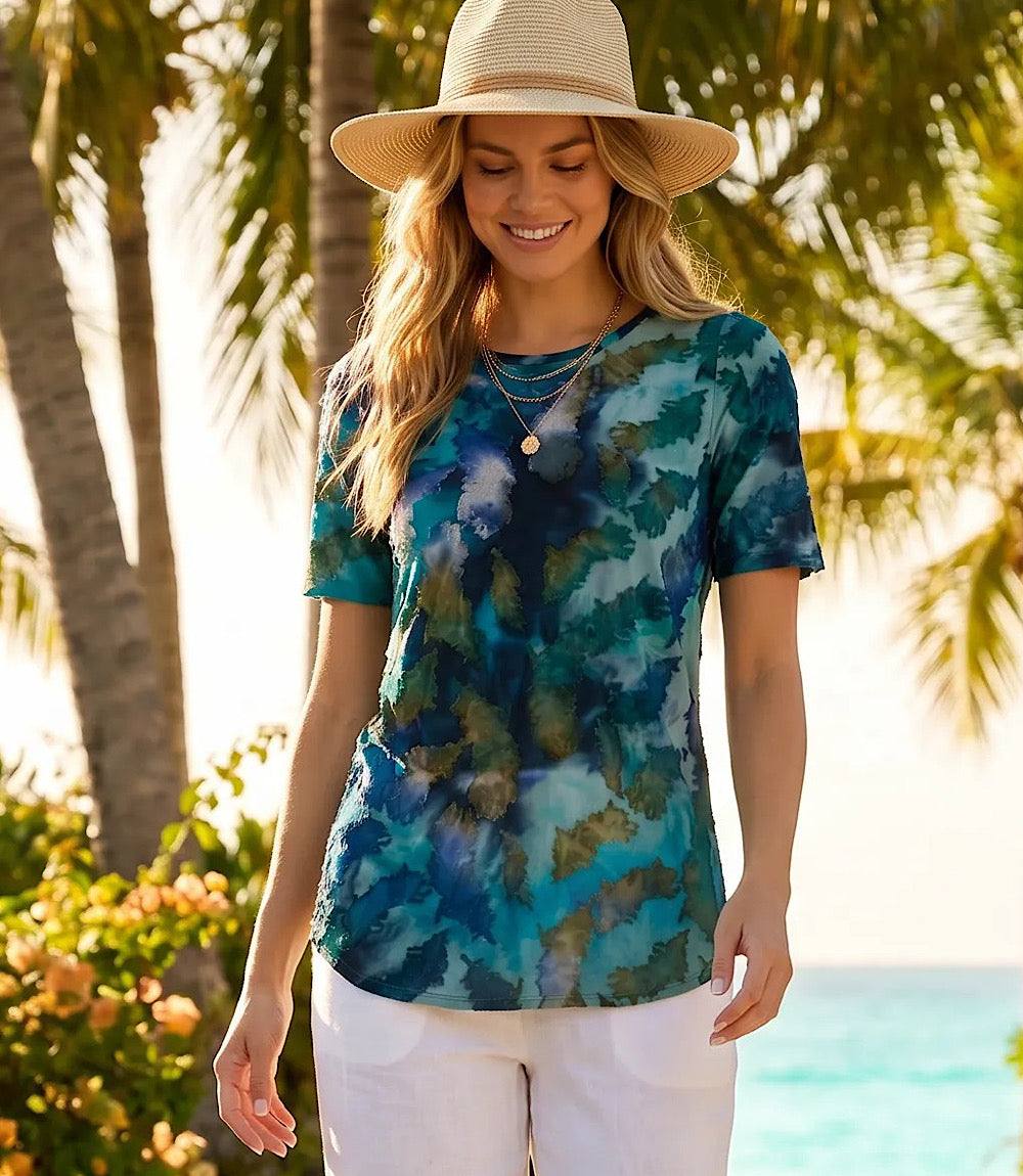 Woman wearing a colorful tie-dye shirt and straw hat in a tropical setting with palm trees and ocean.