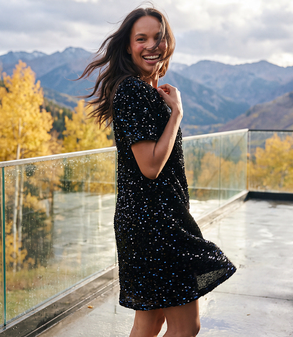 A woman wearing a black short-sleeve dress with blue and white sequins stands on a glass-rail balcony with mountains and autumn trees in the background.