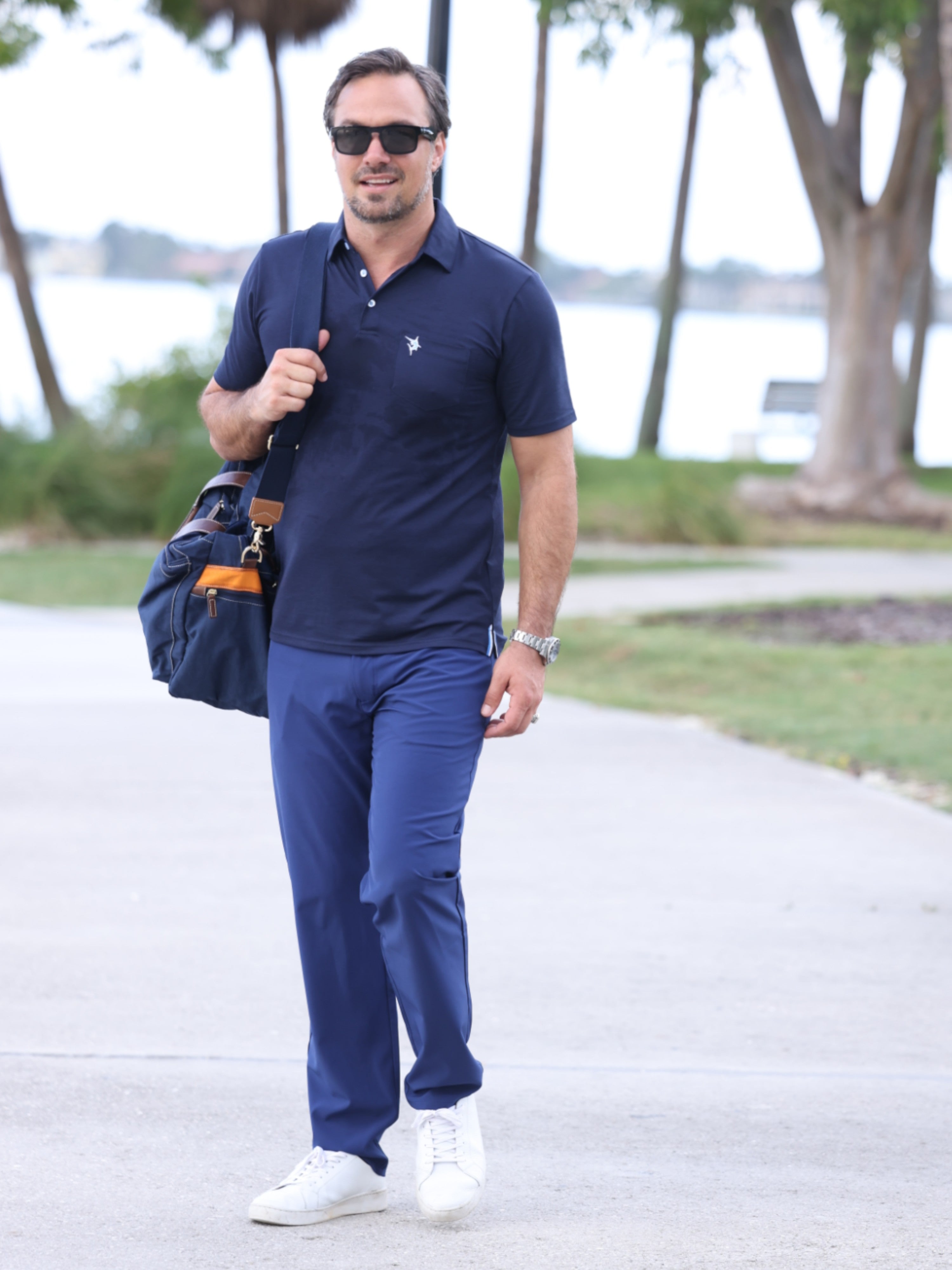 Man in navy polo shirt and pants walking outdoors with a bag.