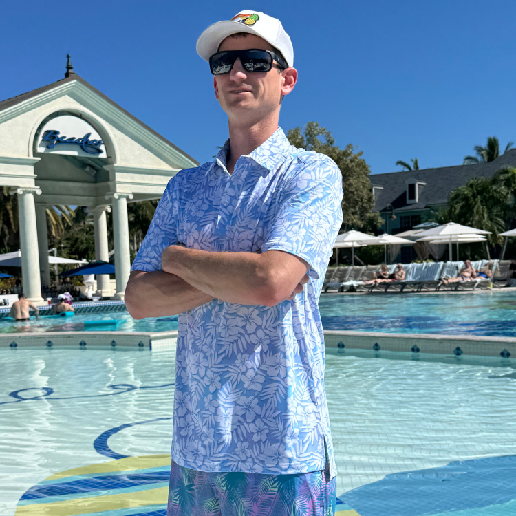 Man in a blue patterned shirt and sunglasses standing by a pool with a building and umbrellas in the background.