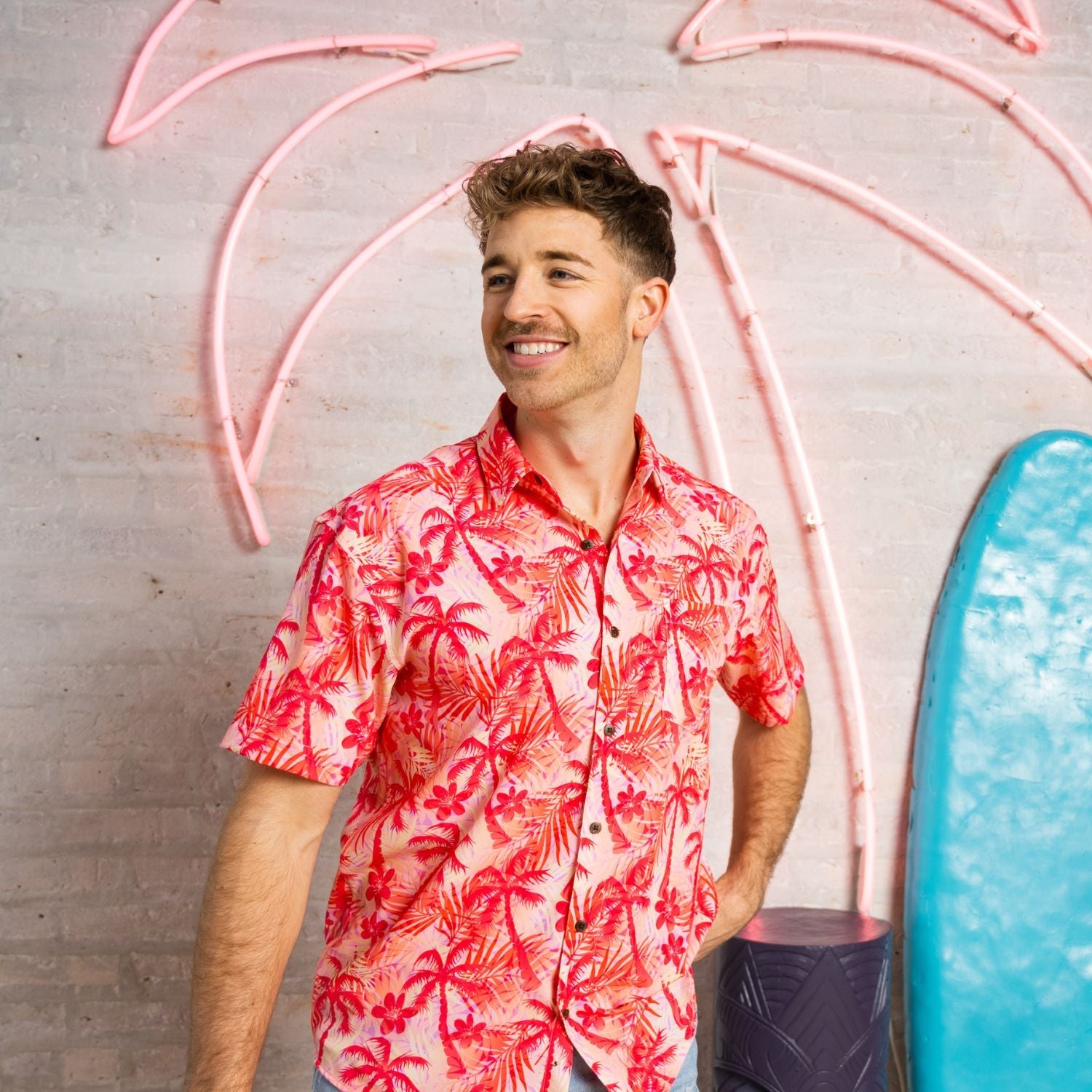 Man wearing a red floral shirt standing in front of pink neon palm trees and a blue surfboard.
